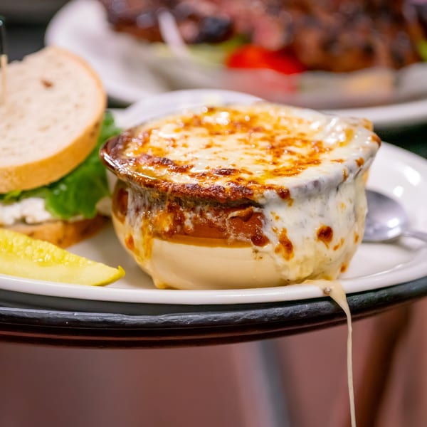 A bowl of P&G's French onion soup with melted cheese, a sandwich, and a pickle.