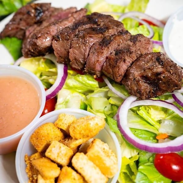 Grilled steak slices on a bed of fresh green lettuce, red onions, and cherry tomatoes, with croutons and dressing.