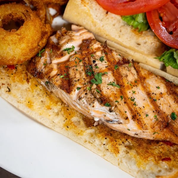 Grilled salmon sandwich with lettuce, tomato, and a side of crispy onion rings from P&G's.